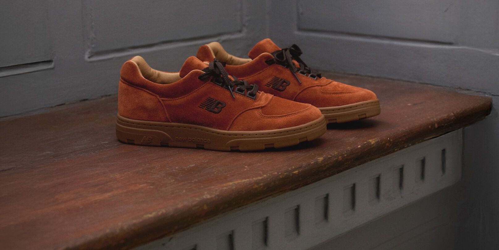 A pair of New Balance Made in UK sneakers sitting on a wooden window seat with panel detailing on the walls.   